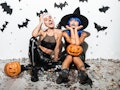 Two pretty young women in leather halloween costumes posing with curved pumpkins over bats and confe...