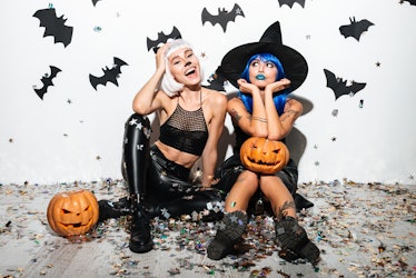 Two pretty young women in leather halloween costumes posing with curved pumpkins over bats and confe...