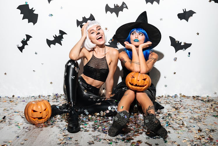 Two pretty young women in leather halloween costumes posing with curved pumpkins over bats and confe...