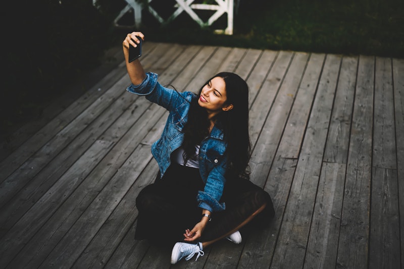 Stylish hipster girl with gorgeous long brunette hair making selfie photos on modern cellular sittin...