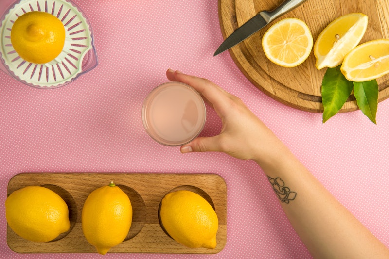 top view of female hand with glass of lemon juice on pink table with squeezer and lemons on wooden b...