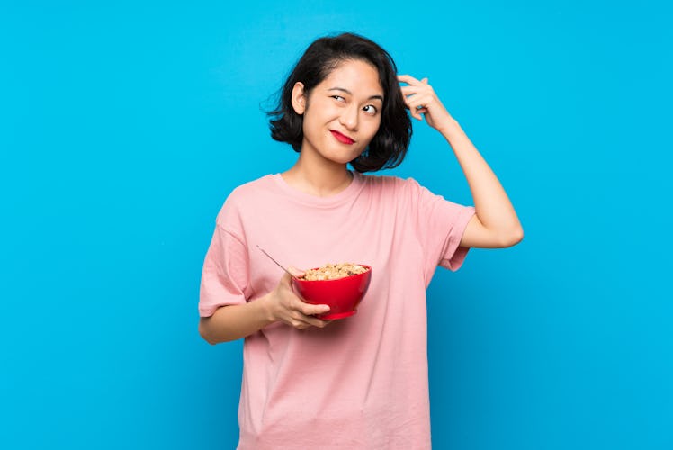 Asian young woman holding a bowl of cereals having doubts and with confuse face expression