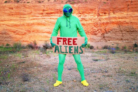 a man dressed in a green alien carnival costume suit at a lone rally with a sign "free aliens" on th...