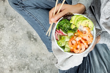 Girl in jeans holding shrimp poke bowl with seaweed, avocado, cucumber, radish, sesame seeds. 