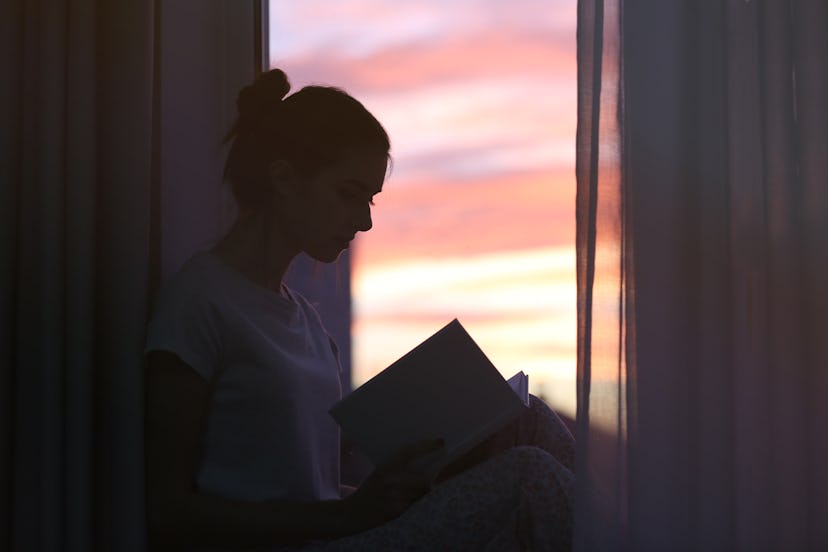 Silhouette of young woman reading book on windowsill at home