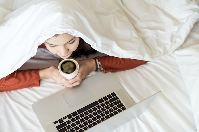 Charming young woman covering head with white blanket lying on bed and watching tv or movies streami...