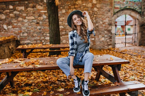 Carefree young woman in trendy vintage pants sitting on table in park and laughing. Curly cute girl ...