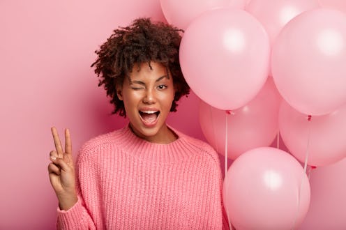 Dark skinned female model blinks eye, shows peace gesture, wears pink jumper and carries air balloon...
