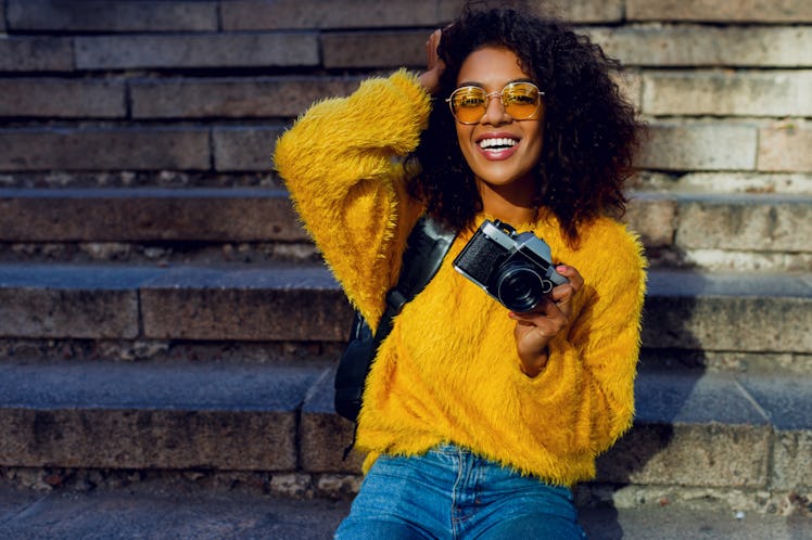 Portrait of  fabulous American student girl with curly dark hair with retro camera sitting on stairs...