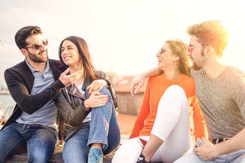 Two couples of friends talking and relaxing together sitting in a small wall during the sunset - mid...