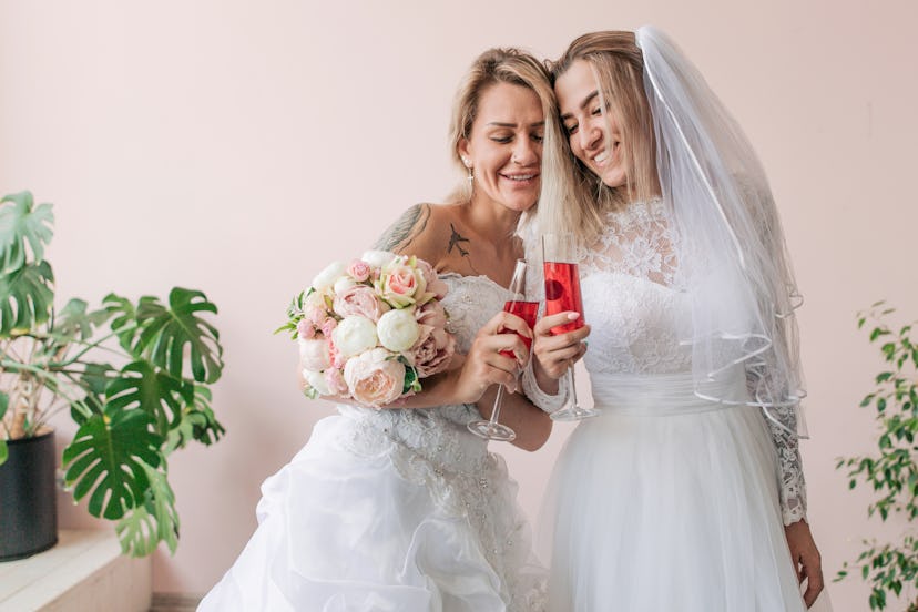 Lesbian in wedding dresses holding bouquet of flowers with champagne standing on pink wall backgroun...