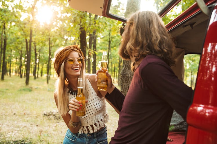 Photo of cheerful hippie couple man and woman smiling and drinking beer in forest near retro minivan
