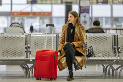 A woman in a long brown winter coat and boots with a red suitcase sits at the airport.