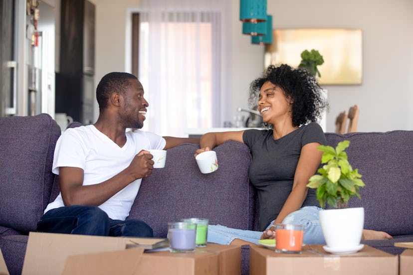 Happy afro couple rest on sofa in living room talking planning future life holds cups drinking tea, ...