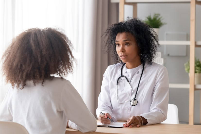Friendly African American pediatrician doctor in white uniform with stethoscope talking to teen girl...