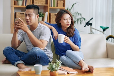Asian couple sitting on couch and man surfing smartphone while woman looking bored and?lonely