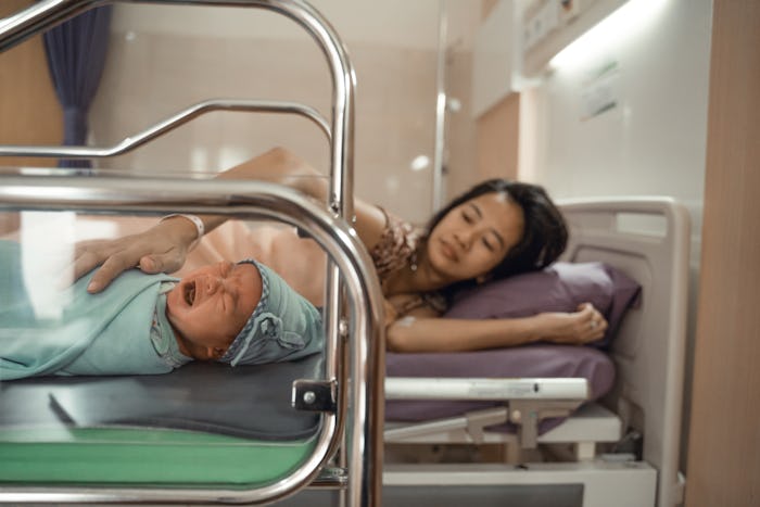 Crying newborn baby laying in crib with his mother lying on bed side in the hospital