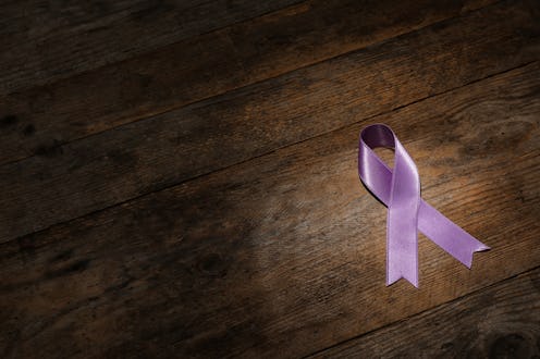 Purple ribbon on brown wooden background, space for text. Domestic violence awareness