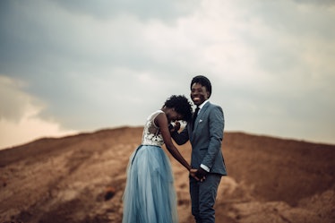 Black happy newlyweds hold hands, cheerful laugh and stand against beautiful landscape.