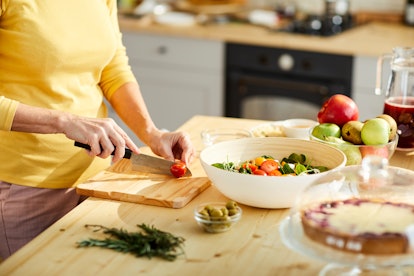 Close-up of unrecognizable woman in yellow sweater standing at kitchen counter and cutting tomato wh...