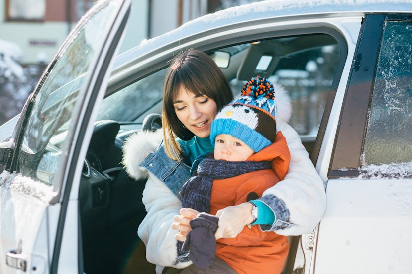 Happy family concept. Young mother on driver's seat of car and trying to wear mittens her cute toddl...