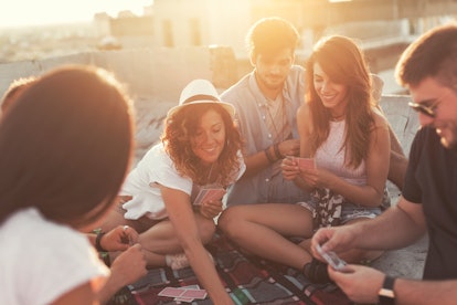 Group of young people sitting on a picnic blanket, having fun while playing cards on the building ro...