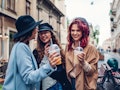 Three stylish women drink out of to-go cups and laugh while hanging out outside a café on Thanksgivi...