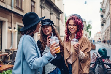 Three stylish women drink out of to-go cups and laugh while hanging out outside a café on Thanksgivi...