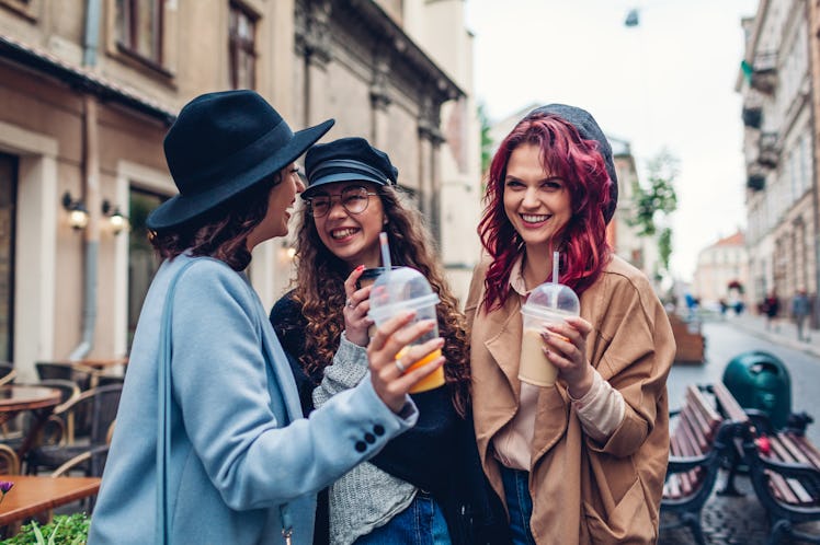 Three stylish women drink out of to-go cups and laugh while hanging out outside a café on Thanksgivi...