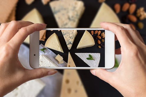 Woman hands taking a photo of different kinds of cheeses on black stone board with nuts and herbs.