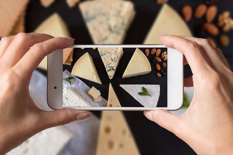 Woman hands taking a photo of different kinds of cheeses on black stone board with nuts and herbs.