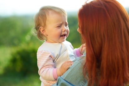 Crying daughter sitting on mother's hands during sunny day in the field. Young mom trying to calm do...