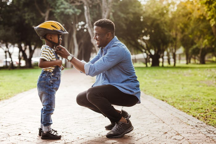 African man putting helmet on cute boy at the park. Father puts his son a protective helmet for ridi...