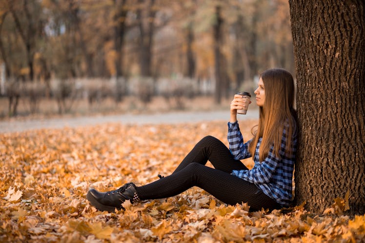 Fall concept. Happy and cheerful woman drinking coffee while sitting on park leaves under fall folia...