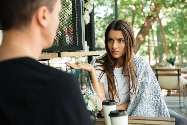 Confused young woman gesturing with hand and looking at her boyfriend while sitting in a cafe at the...