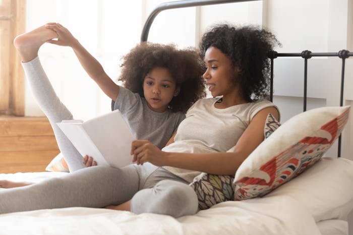 African american family mom and cute little kid daughter read book fairy tale sitting in bed, small ...