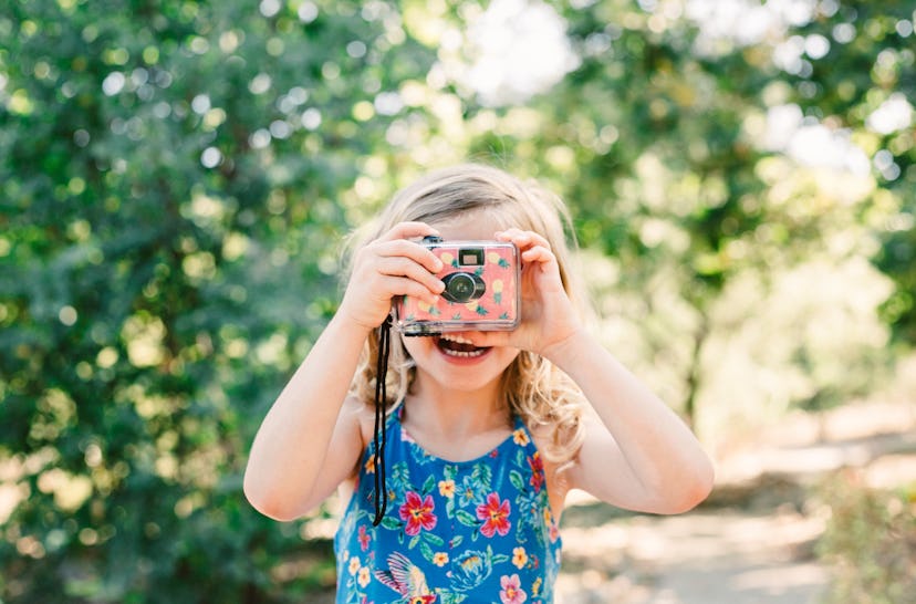 A little girl with blonde curly hair take a picture with a disposable camera decorated with pineappl...