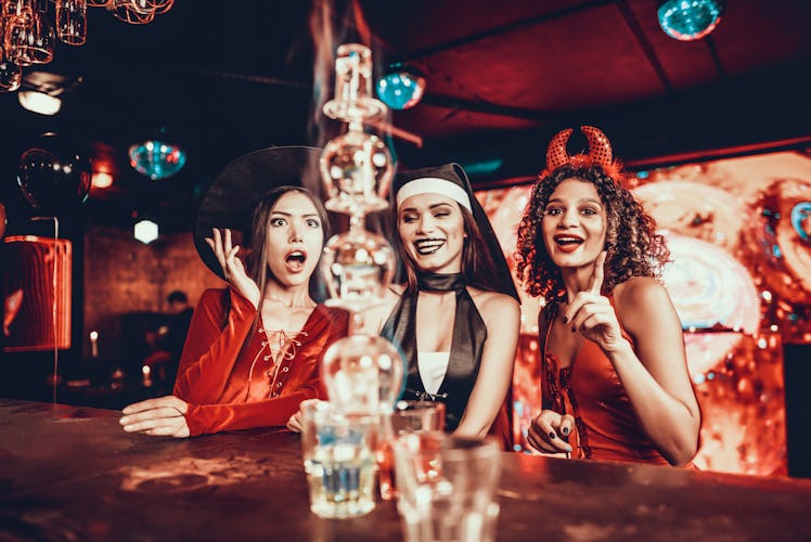 Women in Costumes Looking at Flaming Cocktail. Group of Smiling Young Friends Wearing Costumes Excit...