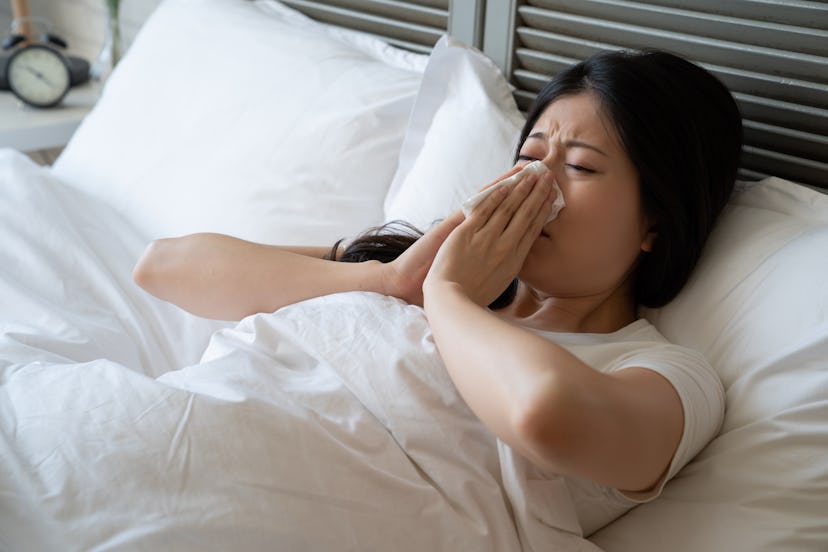 sick asian woman suffering from cold lying in bed with tissue sneezing. ill chinese female cover hid...