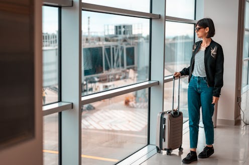 Woman in bomber jacket and jeans waiting at the airport with carry-on luggage.