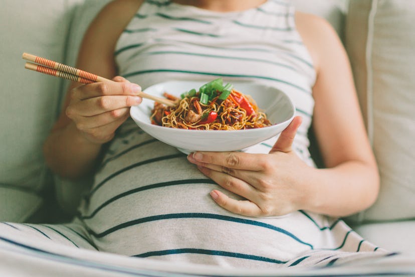 A pregnant woman eating a bowl of noodles, resting on her belly.