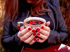 Autumn. Girl with bright red nails siting in autumn park and holding cup of tea with dogrose berries...