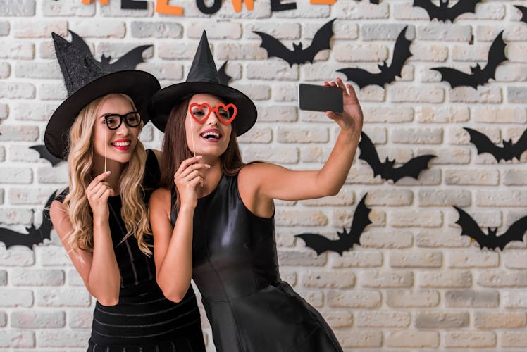 Beautiful girls in black dresses and witch hats are holding party props and doing selfie using a sma...