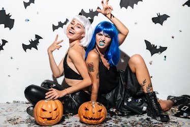 Two cheerful young women in leather halloween costumes posing with curved pumpkins over bats and con...
