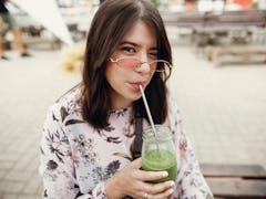 Stylish hipster boho girl drinking spinach smoothie in a glass jar with metal reusable straw at a st...