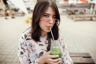 Stylish hipster boho girl drinking spinach smoothie in a glass jar with metal reusable straw at a st...
