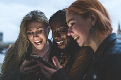 Best friends girls with smartphones laughing. Three women in London at dusk, looking at phones and l...