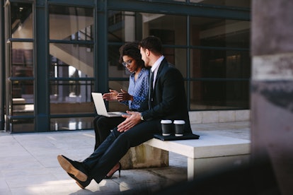 Two business people sitting on a bench in modern office building with a laptop. Woman with laptop di...