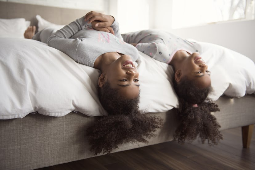 The Two smiling little african american girls on bed at home heads leaning over side of bed 