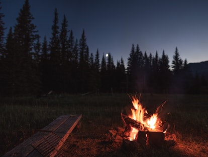 Campfire under the out of focus moon. There is a wooden bench beside the campfire with a hot dog / m...